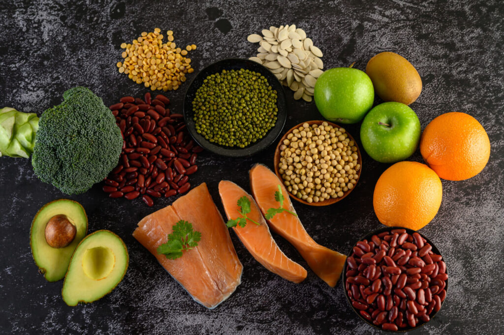 Various healthy foods, including legumes, broccoli, fruits, and salmon, artfully arranged on a black cement floor.