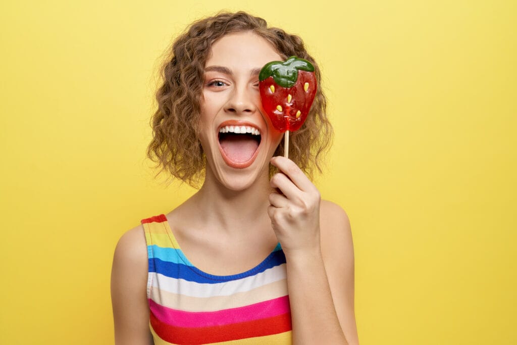 Happy, cheerful girl holding sweet red strawberry lollipop and having fun. Playful model in striped dress looking at camera, posing on yellow background.