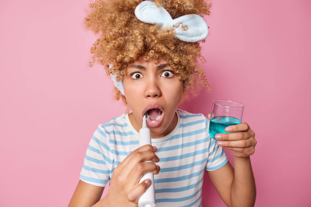 Shocked young woman brushes teeth with electric brush, holds glass of blue mouthwash - daily hygiene concept